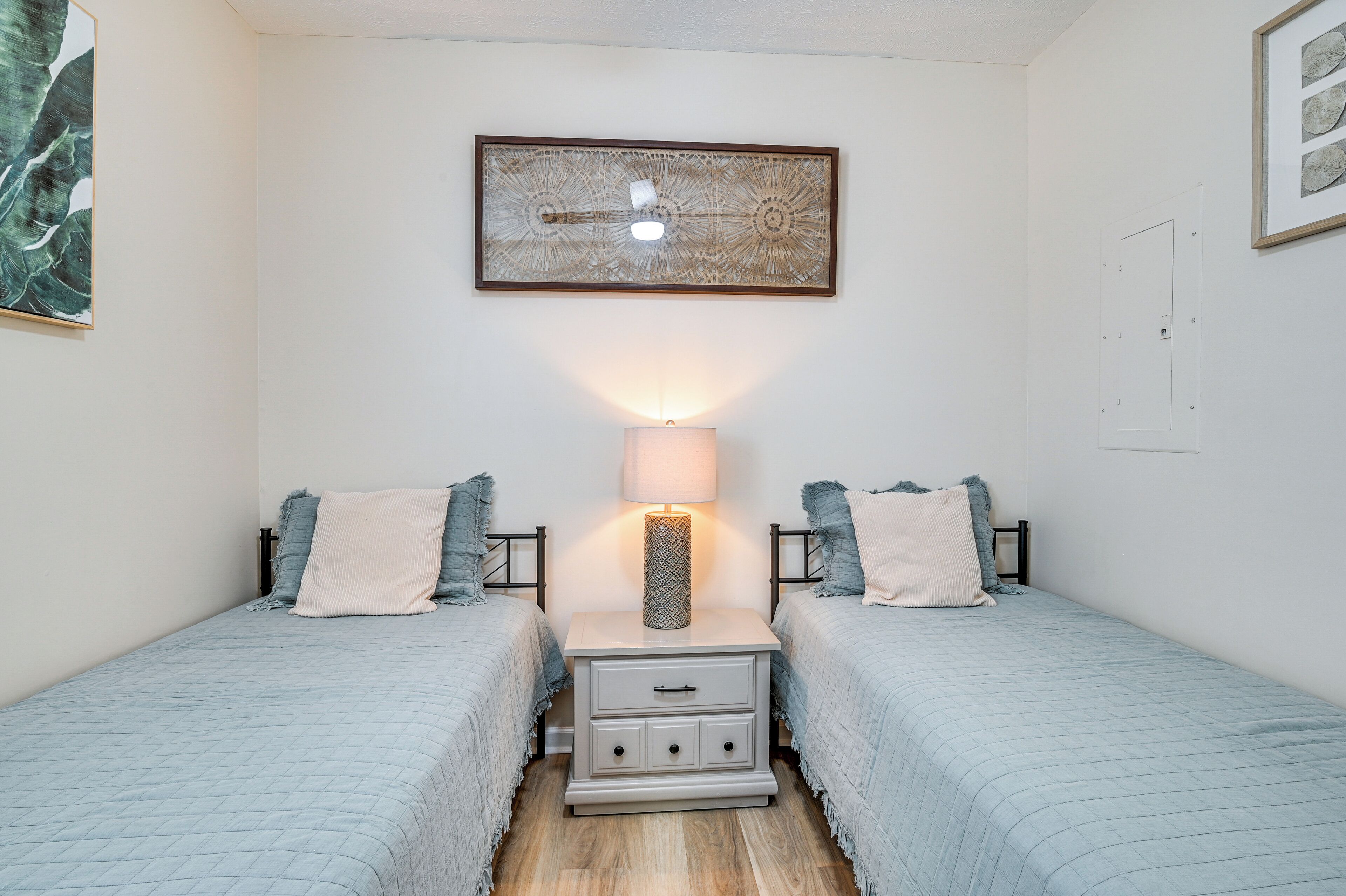 A bedroom with two beds, a bedside table with a lamp, and a framed artwork on the wall.
