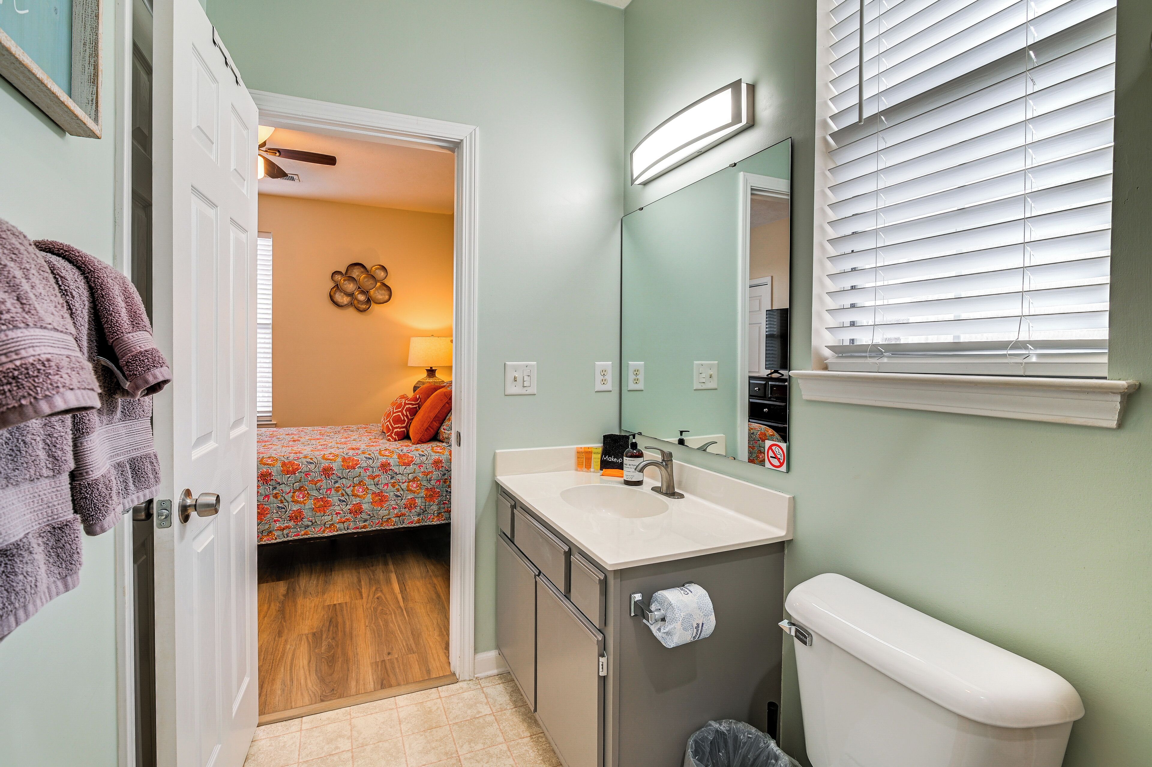 A compact bathroom with a toilet, sink, and mirror. A bed with floral bedding is visible through an open door.
