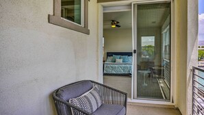Un balcon avec un fauteuil en osier, une porte-fenêtre coulissante donnant sur une chambre à coucher équipée d’un lit et d’un ventilateur de plafond.