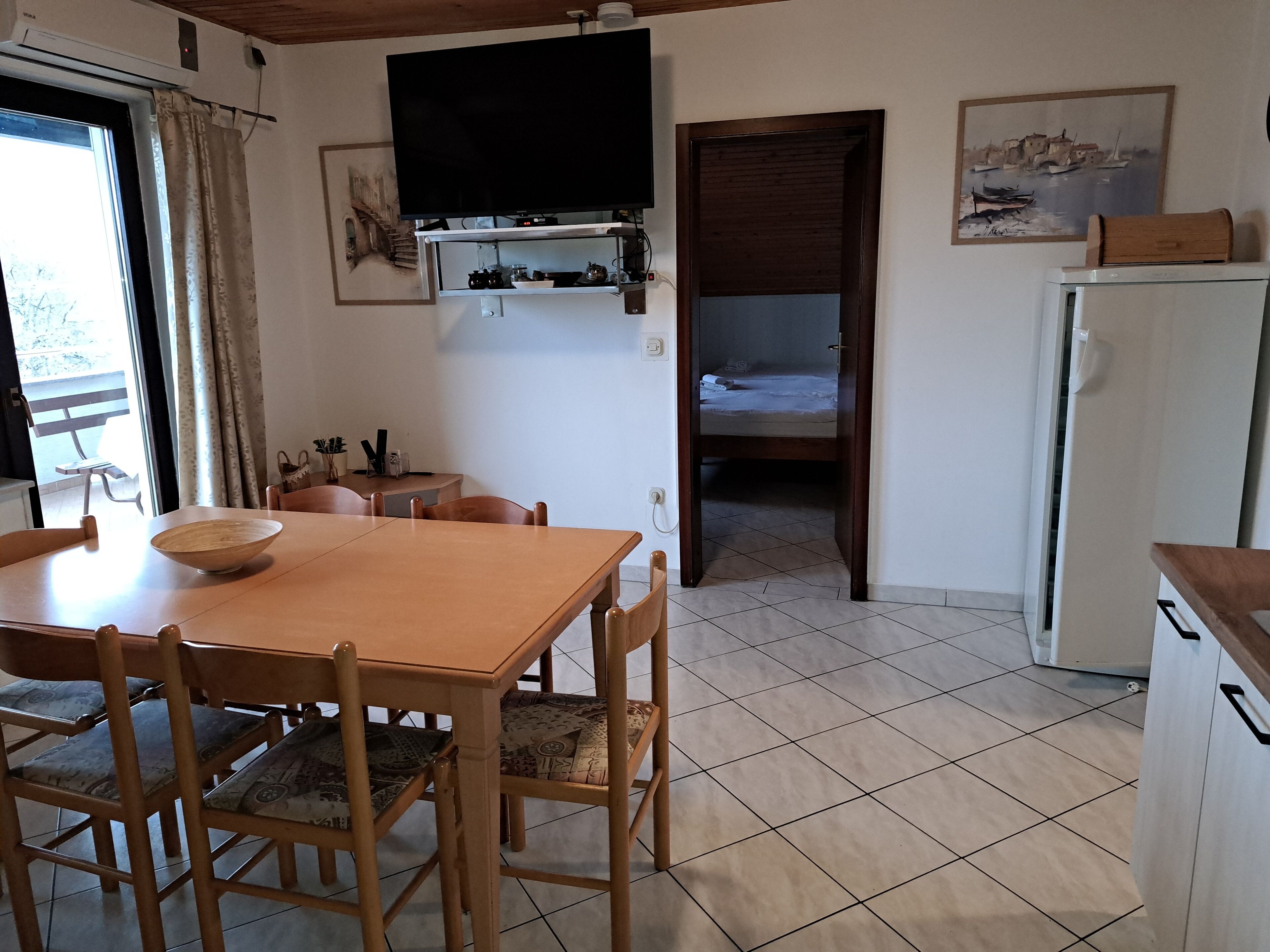 A compact kitchen with a dining area, a TV mounted above the counter, and a bed in the adjacent room.