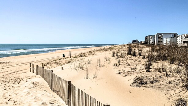 Una playa con una valla de madera, dunas arenosas y un edificio de varios pisos al fondo.