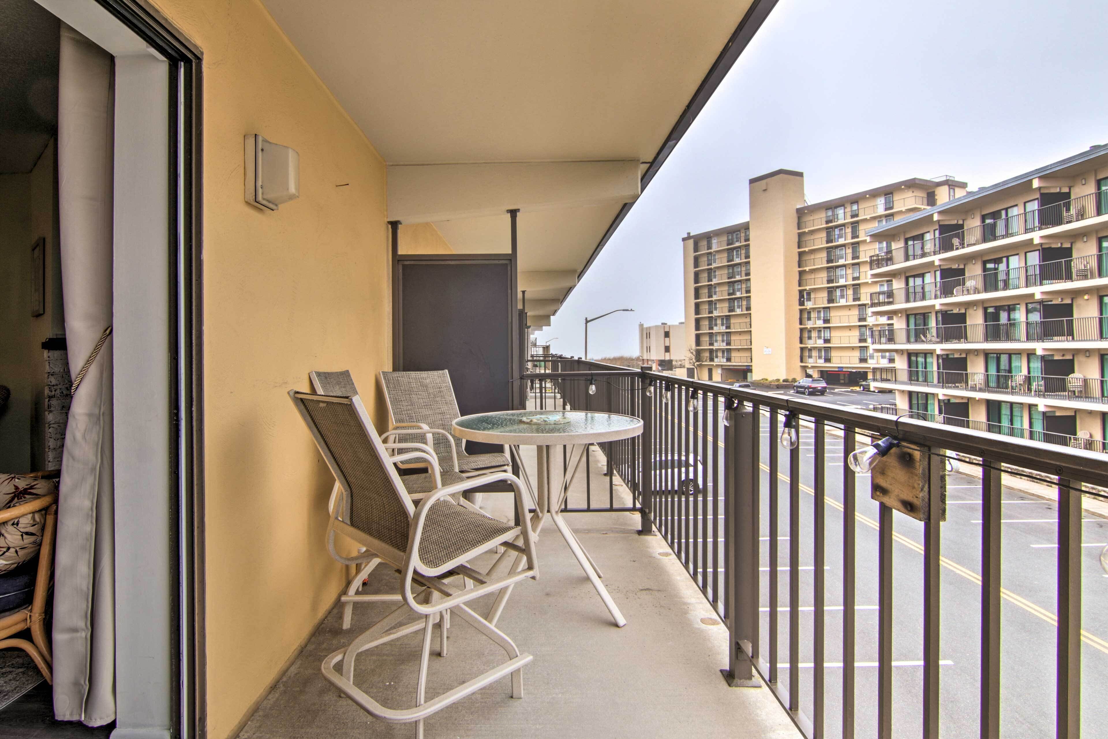 A balcony with wicker chairs and a small table, overlooking a street with buildings in the background.