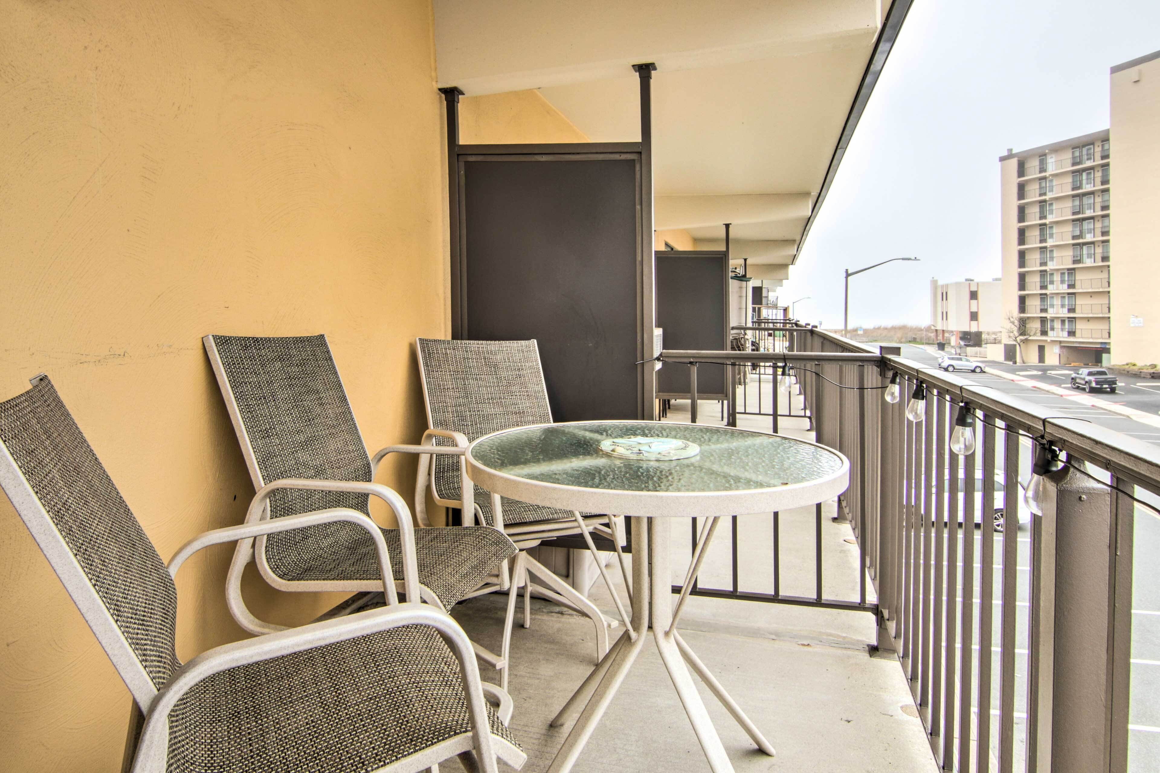 A balcony with a round glass table and two wicker chairs, overlooking a street with buildings and parked cars.