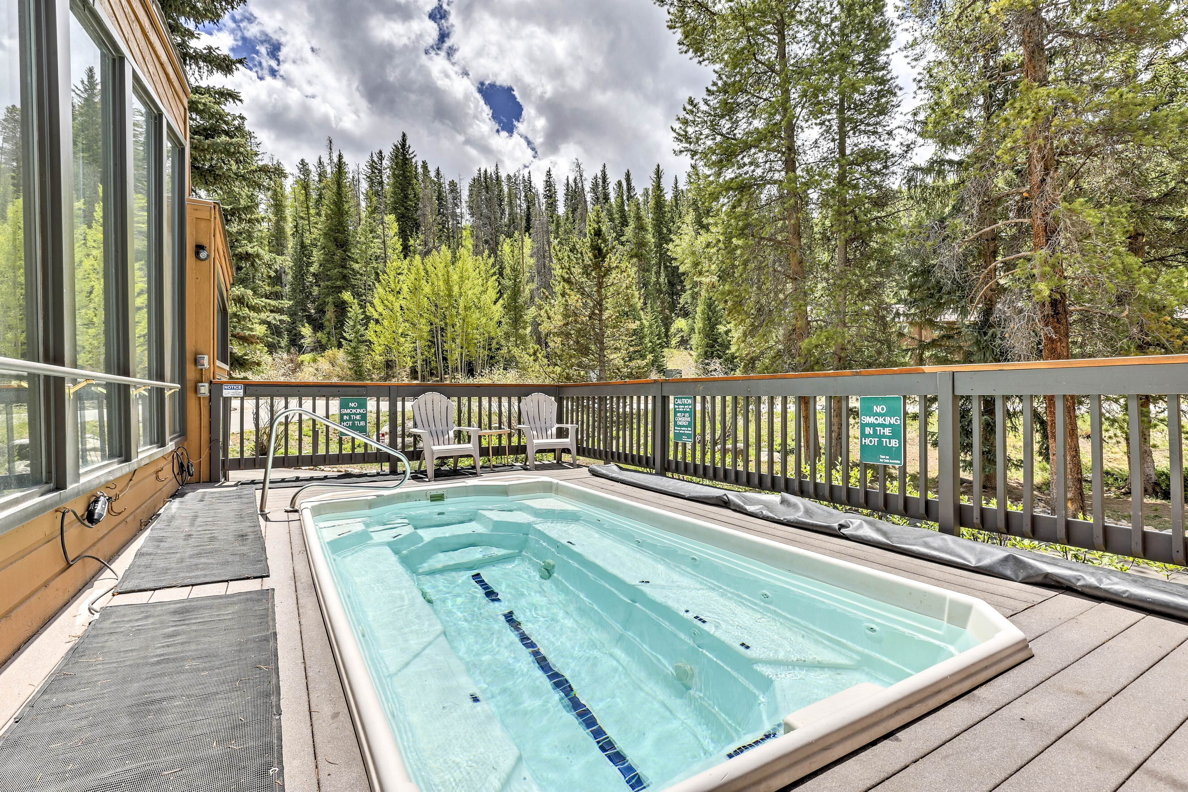 A hot tub surrounded by a wooden deck, with a view of a forested area and a sign prohibiting smoking.