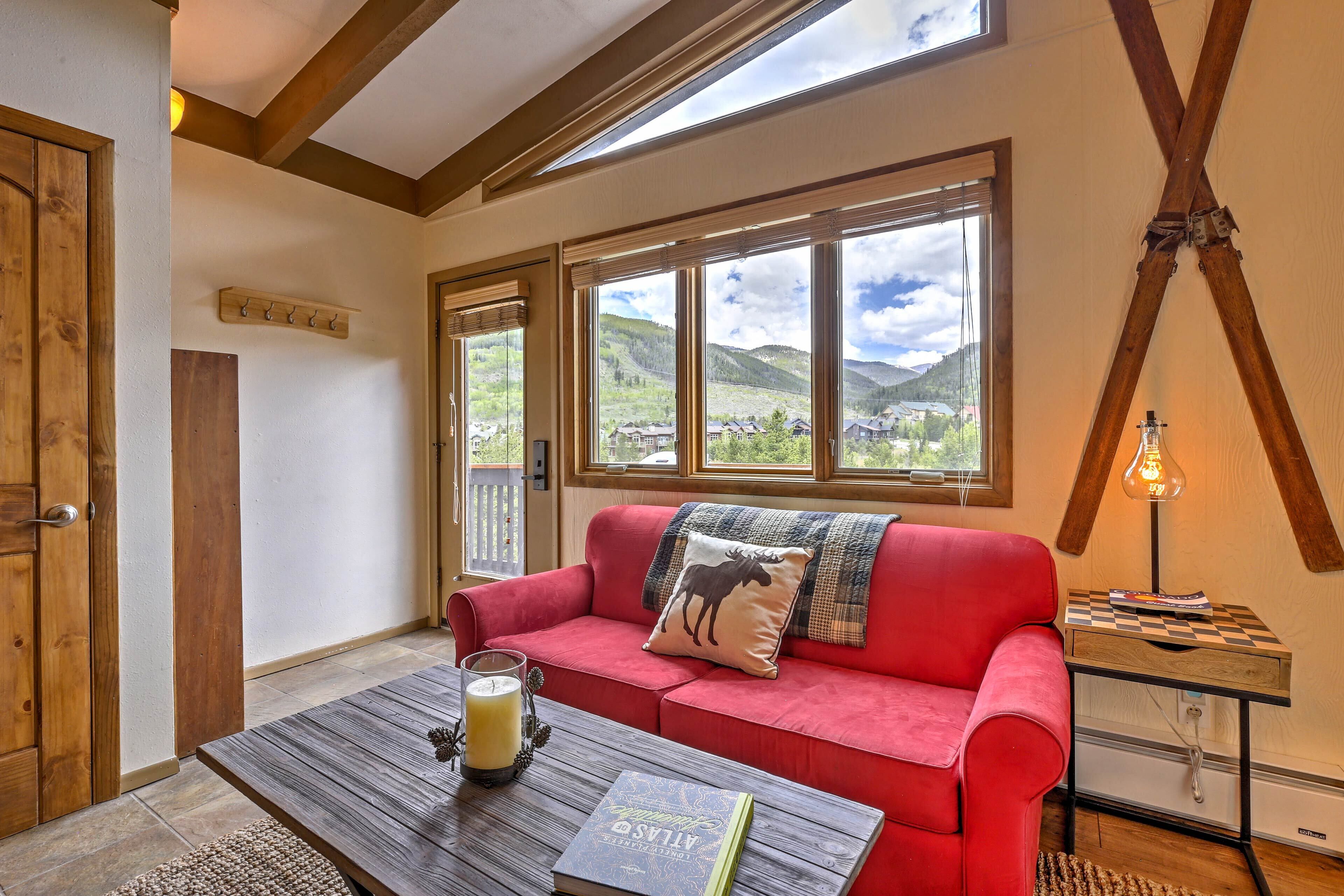 A cozy room with a red sofa, a wooden coffee table with a book, a lamp, and a view of mountains through large windows.