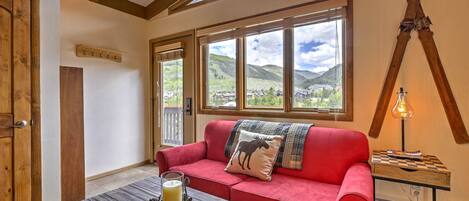 A cozy room with a red sofa, a wooden coffee table with a book, a lamp, and a view of mountains through large windows.