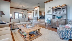 A spacious living room with a wooden coffee table, a sofa, and a patterned armchair. There is a kitchen in the background with cabinets and a dining area. - Serene Hill Country Wildlife Sanctuary w/ Porch (Boerne)