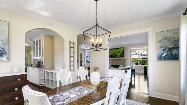 A dining area with a wooden table, white chairs, and a chandelier. There is a kitchen in the background with white cabinets and a stone backsplash. - Charming Old Northwood Home ~ 3 Mi to Beach! (West Palm Beach)