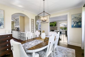 A dining area with a wooden table, white chairs, and a chandelier. There is a kitchen in the background with white cabinets and a stone backsplash.