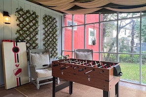 A cozy room with a wooden foosball table, two white armchairs, a red wall, and a large window with a view of a house and greenery. - Charming Emerald Lakes Cottage w/ Hot Tub! (Long Pond)
