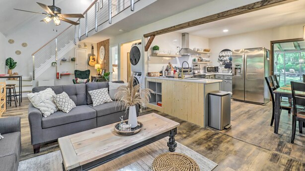 A spacious living area with a grey sofa, wooden coffee table, and a ceiling fan. A kitchen with modern appliances and a dining area with a table and chairs is visible in the background. - Charming Emerald Lakes Cottage w/ Hot Tub! (Long Pond)