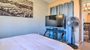 A bedroom with a bed, a flat-screen TV, a standing fan, and a painting on the wall. - Cottage With Fire Pit + Deck on The Klamath River! (Hornbrook)