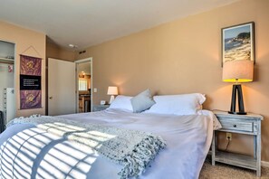Una cama bien hecha con sábanas blancas y un plaid, una lámpara de noche y un cuadro con una playa en la pared.