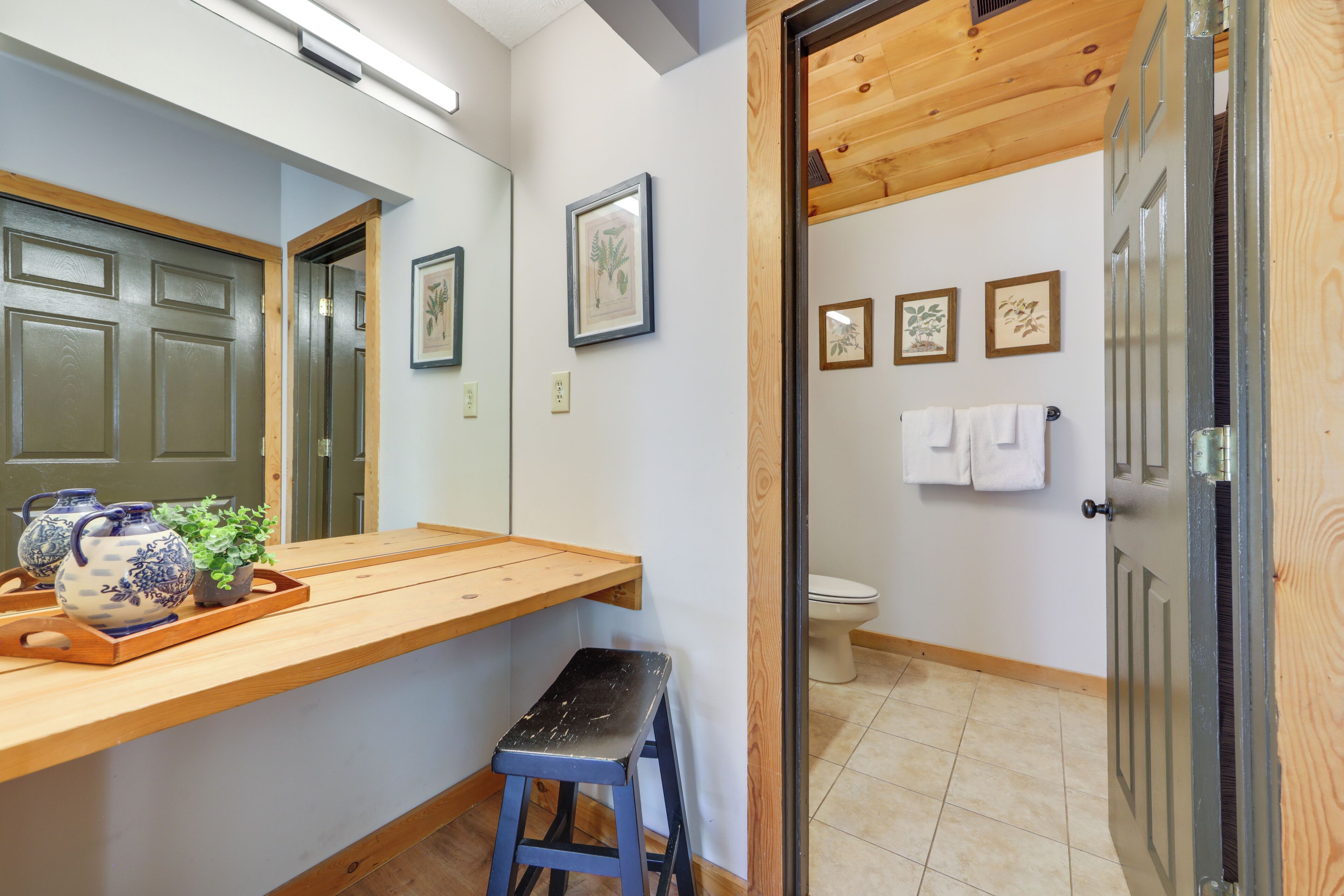 Un baño con tocador de madera, un taburete, un inodoro e impresiones enmarcadas de motivos botánicos en la pared.