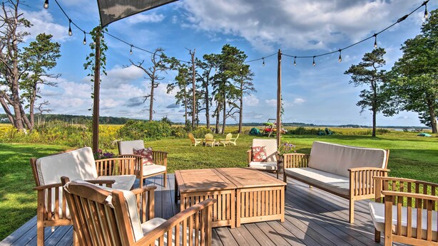 Eine Holzterrasse mit Gartenmöbeln, Lichterketten und Blick auf ein Wiesenfeld und Bäume.