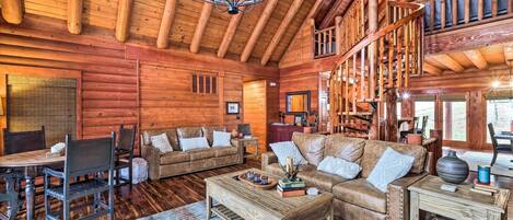 Un intérieur spacieux de chalet en bois, avec une table à manger en bois, des canapés en cuir et un escalier menant à un niveau supérieur.