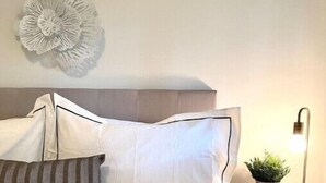 A neatly made bed with white bedding and striped pillows, a decorative wall piece, and a bedside lamp with a plant.