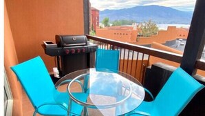 A balcony with a glass table and turquoise chairs, overlooking a mountainous landscape.