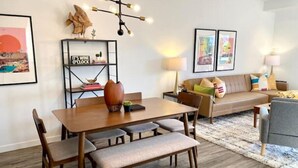 A modern living room with a wooden dining table, cushioned chairs, a sofa, and a coffee table. There is a bookshelf with books and decorative items, a lamp, and framed pictures on the wall.