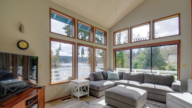 A spacious living room with a large sectional sofa, a flat-screen TV on a wooden stand, and multiple large windows offering views of a lake and trees. - The Swedish Fish: Getaway on Lake Saint Clair! (Olympia)