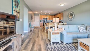Ein Wohnzimmer mit Fernseher, Sofa und Couchtisch. Im Hintergrund ist eine Küche mit Holzschränken und einem Kühlschrank zu sehen.