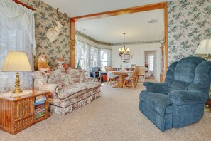 A living room with a floral-patterned sofa, a blue armchair, a wooden side table with books, and a mounted deer head on the wall.