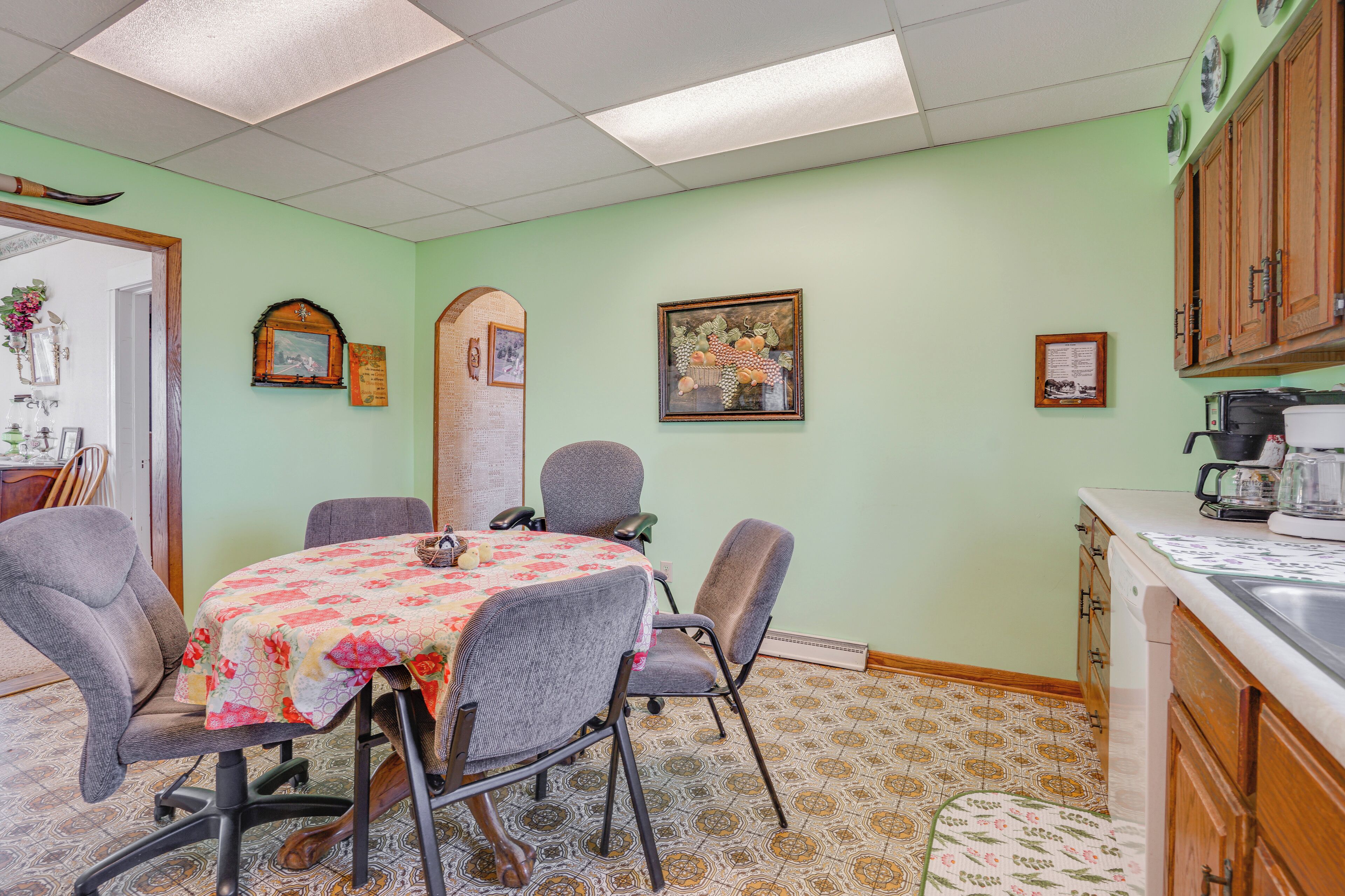 Un comedor con una mesa cubierta con un mantel floral, rodeado de sillas. Hay una zona de cocina con armarios de madera y una cafetera en la encimera.
