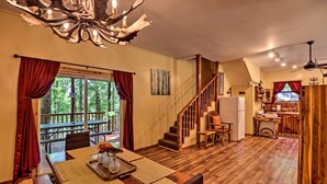 A spacious living area with a wooden floor, a dining table with placemats, a chandelier, and a view of the outdoors through large windows. - Ouachita River Cabin: Royal Haven w/ Dock! (Royal)