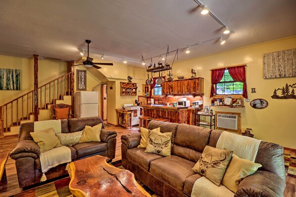 A cozy living room with a fireplace, wooden furniture, and a kitchen area in the background. - Ouachita River Cabin: Royal Haven w/ Dock! (Royal)