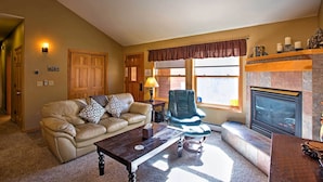 A living room with a beige sofa, a wooden coffee table, a leather armchair, a fireplace, and a window with a brown valance.