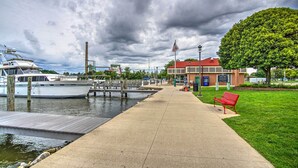 A waterfront walkway with a dock, a boat, a building with a red roof, and a large tree. - Downtown Grand Haven Apt < 1 Mi to Lake Michigan (Grand Haven)