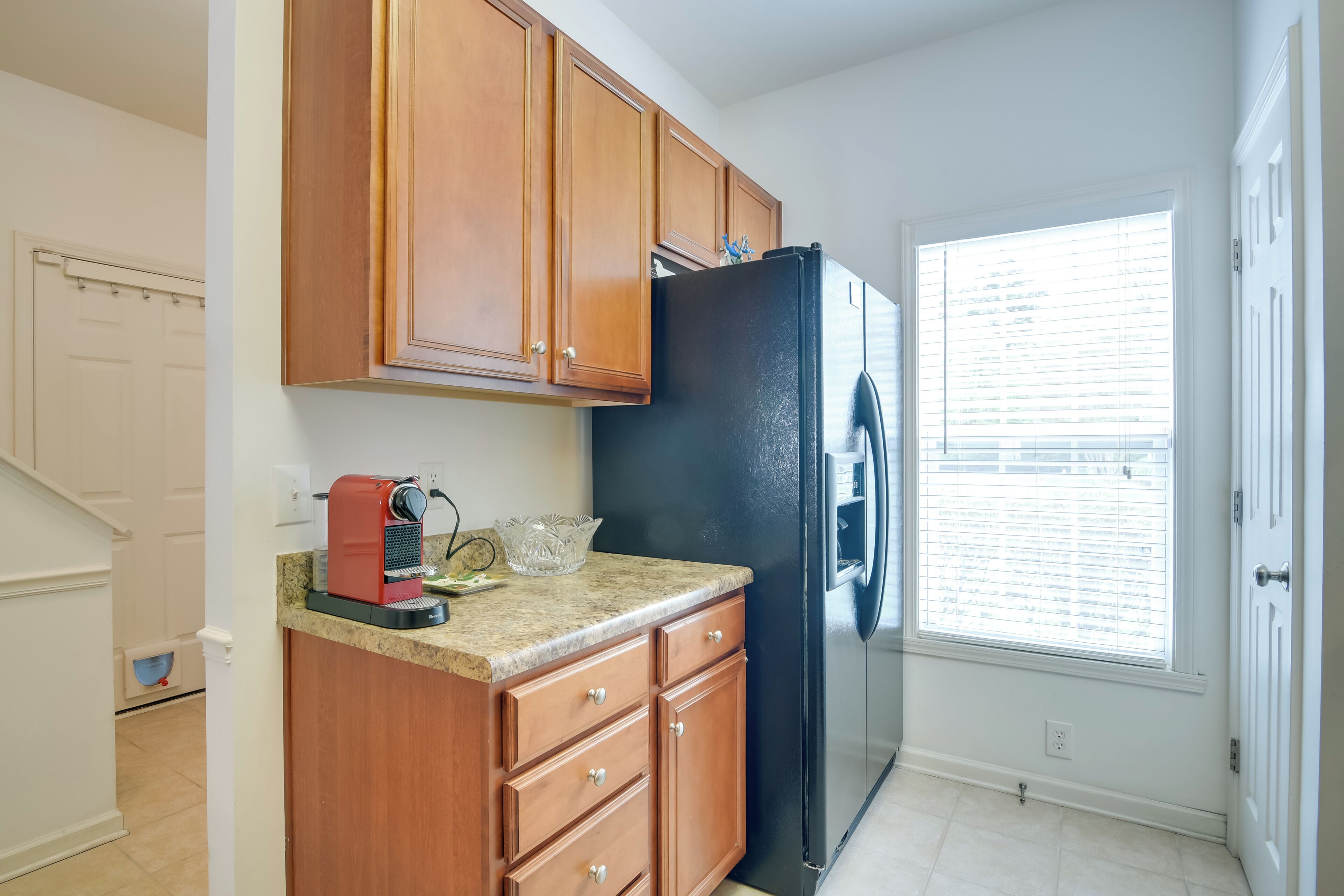 A kitchen with wooden cabinets, a black refrigerator, a coffee maker, and a window with blinds.