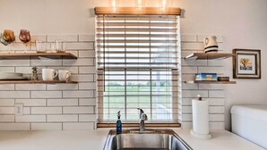 A kitchen with a stainless steel sink, white tiled backsplash, wooden shelves with glassware, and a window with blinds. - On-site Pond & Fire Pit: Montague Retreat! (Montague)
