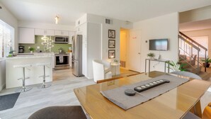 Cocina moderna con armarios blancos, microondas y refrigerador. Hay un comedor con mesa de madera y taburetes de bar. Un televisor está montado en la pared, y una escalera conduce a otro nivel.
