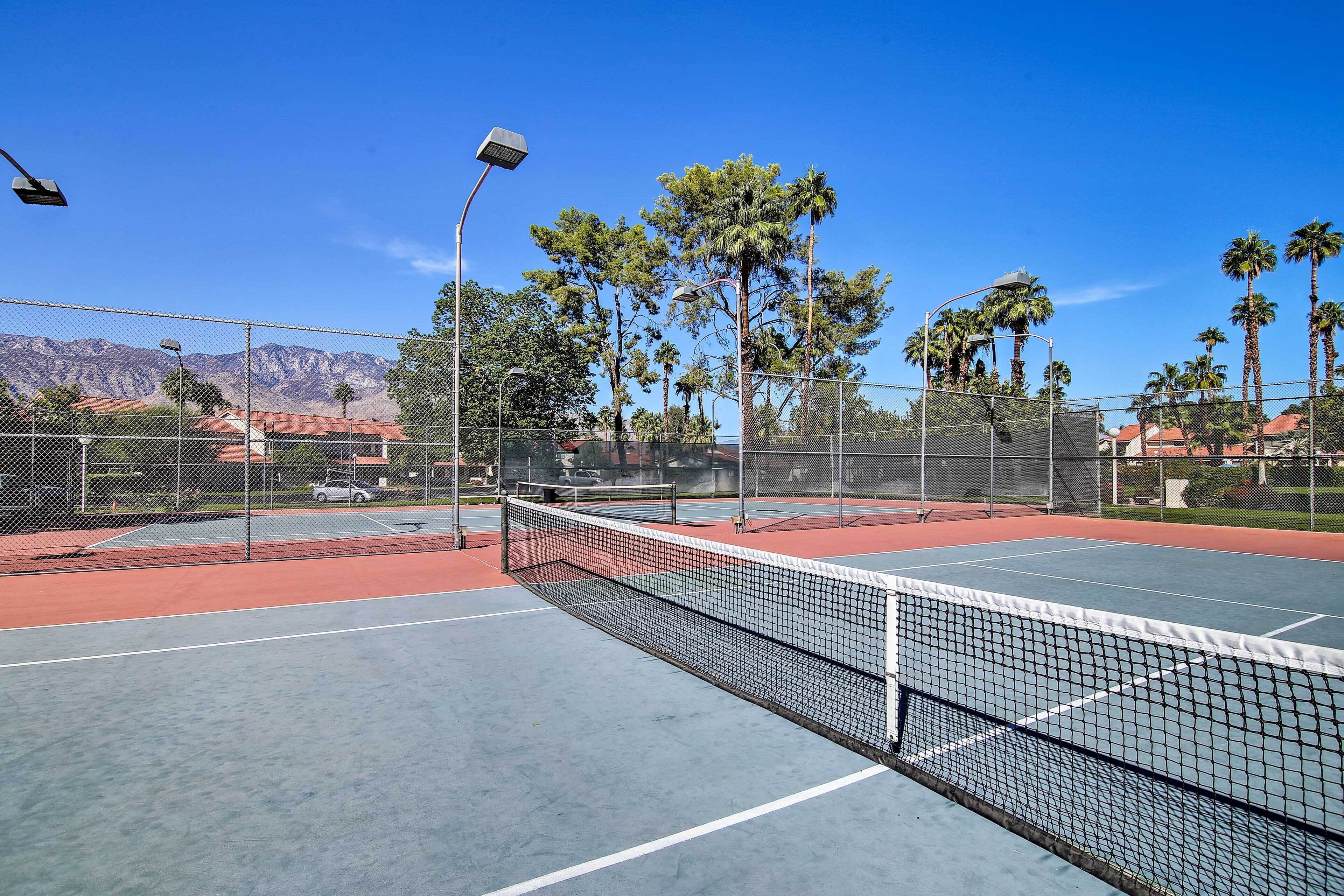 An outdoor tennis court with a net, surrounded by a chain-link fence, palm trees, and a mountain backdrop.
