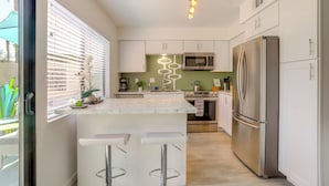 Una cocina moderna con armarios blancos, un refrigerador de acero inoxidable, un microondas y un horno empotrado. Hay dos taburetes de bar en la isla y una ventana con persianas.