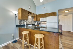 Una cocina con armarios de madera, encimera de granito, dos taburetes de bar, un refrigerador blanco y una lámpara de pie.