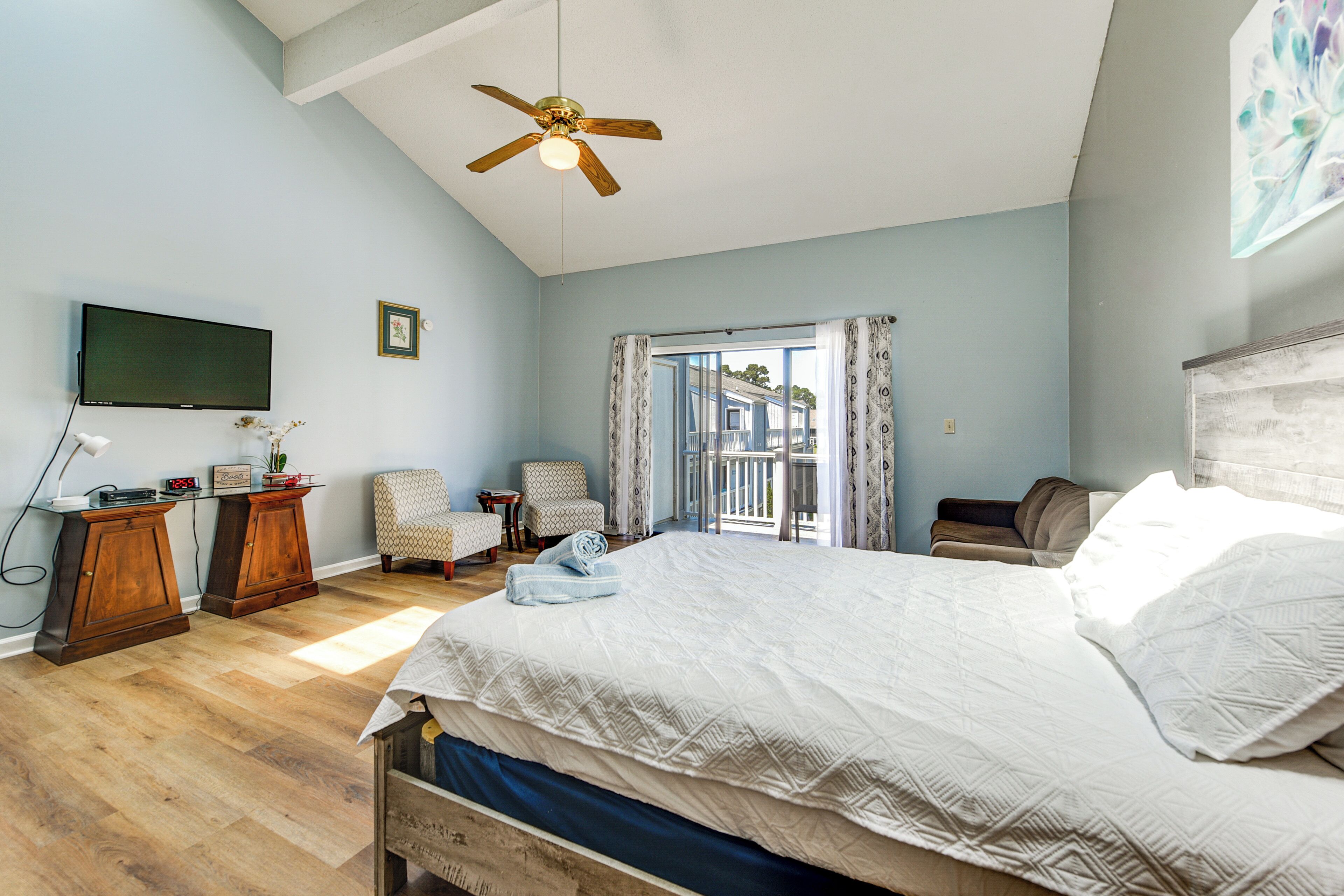 A bedroom with a bed, a ceiling fan, a TV, a chair, a small table, and a window with curtains.