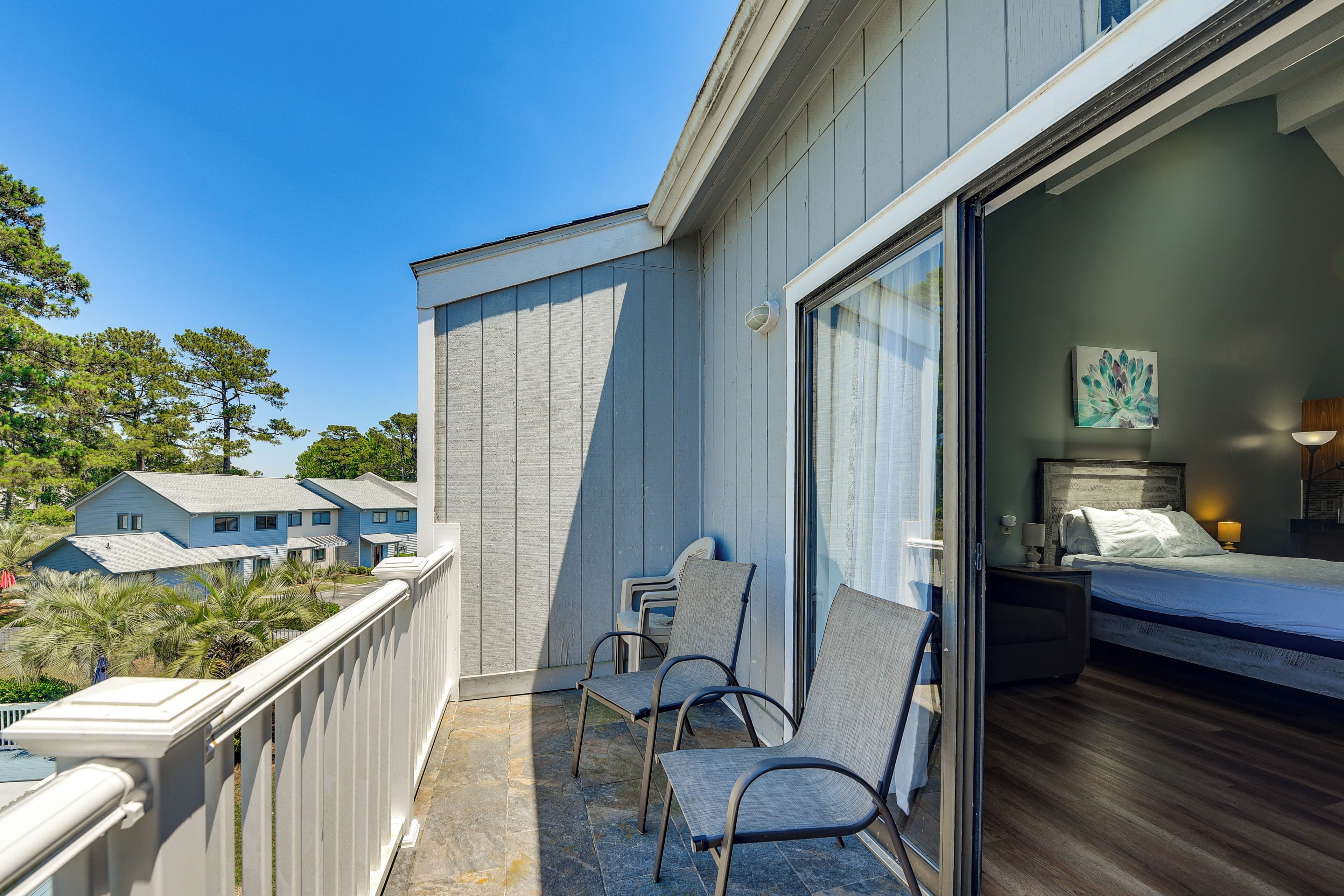 A balcony with chairs, a bed, and a view of neighboring houses.