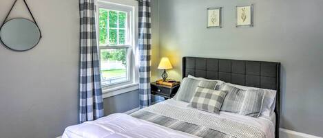 Un dormitorio con cama, una mesita de noche con lámpara, una ventana con cortinas y dos cuadros en la pared.
