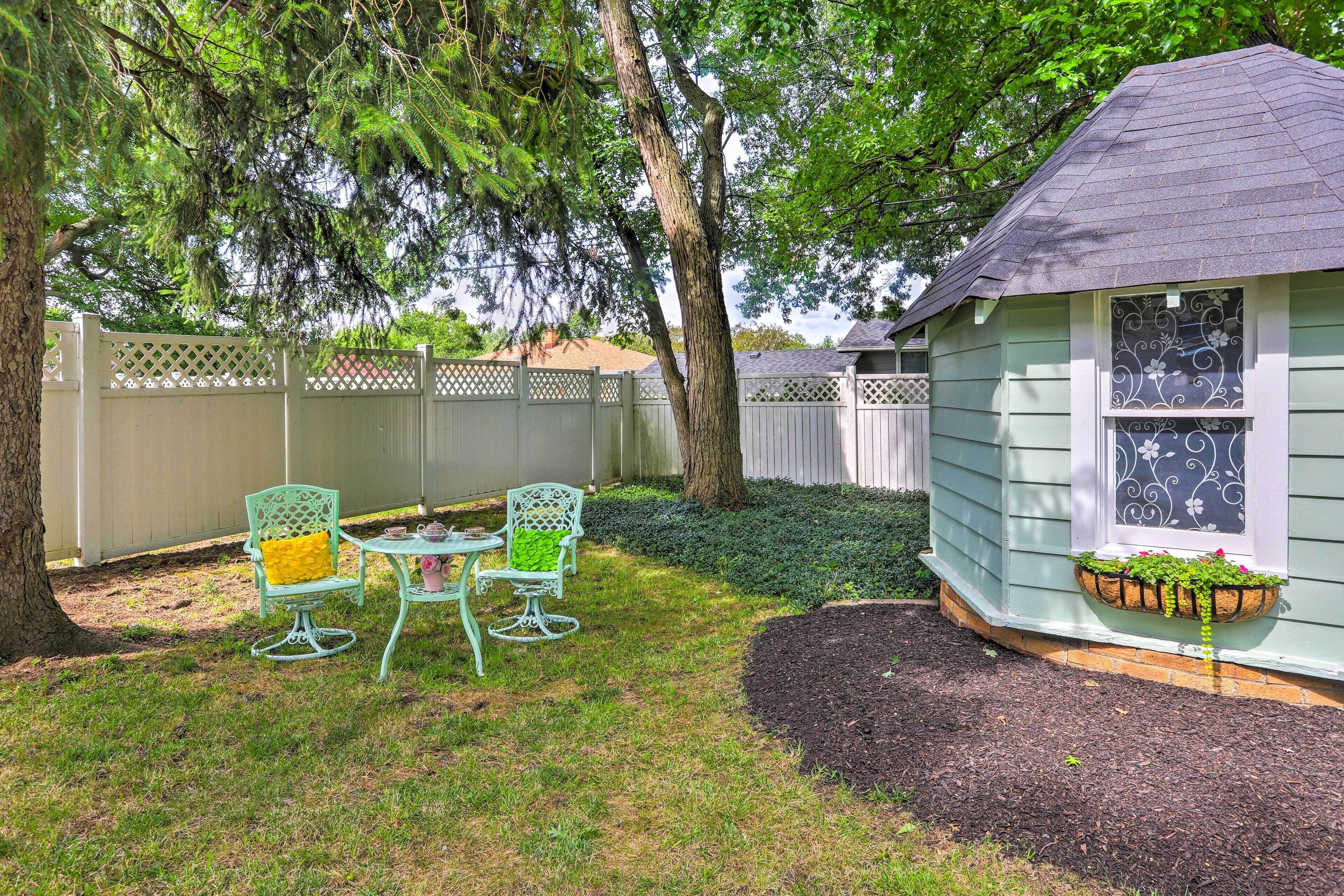 A small, single-story house with a dark roof, a white picket fence, and a garden area with a table and chairs.