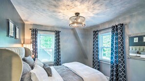 A bedroom with a bed, two windows with patterned curtains, a chandelier, and a ceiling with a textured finish.