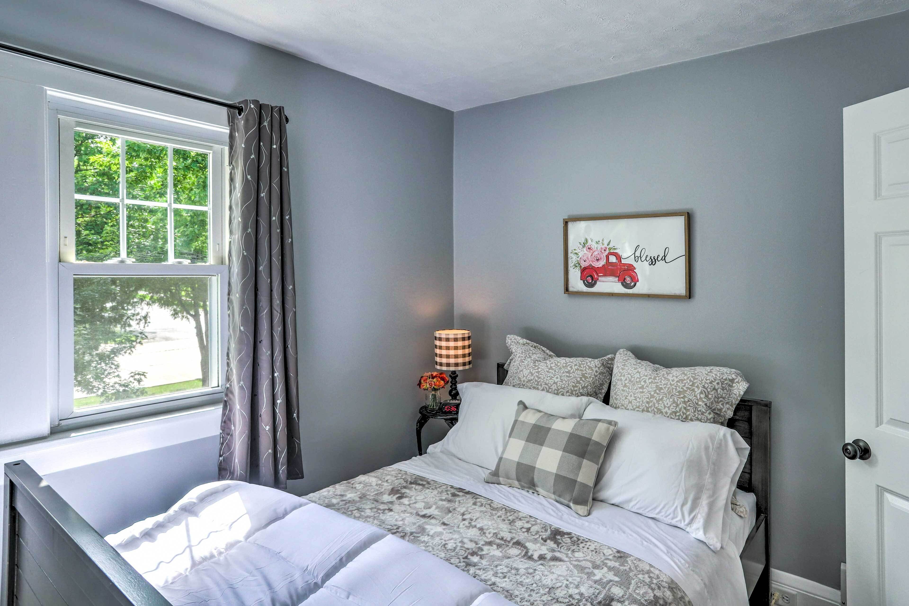 A bedroom with a bed, a window with curtains, a bedside table with a lamp, and a framed picture on the wall.