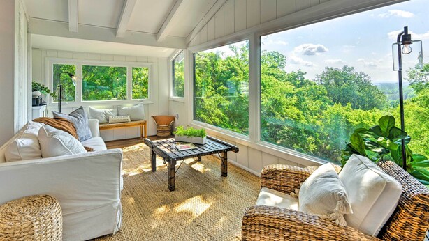 A sunlit room with a large window overlooking a lush green landscape, furnished with a white sofa, wicker chairs, and a wooden coffee table. - Stylish Franklin Cabin on 6 Private Acres! (Franklin)