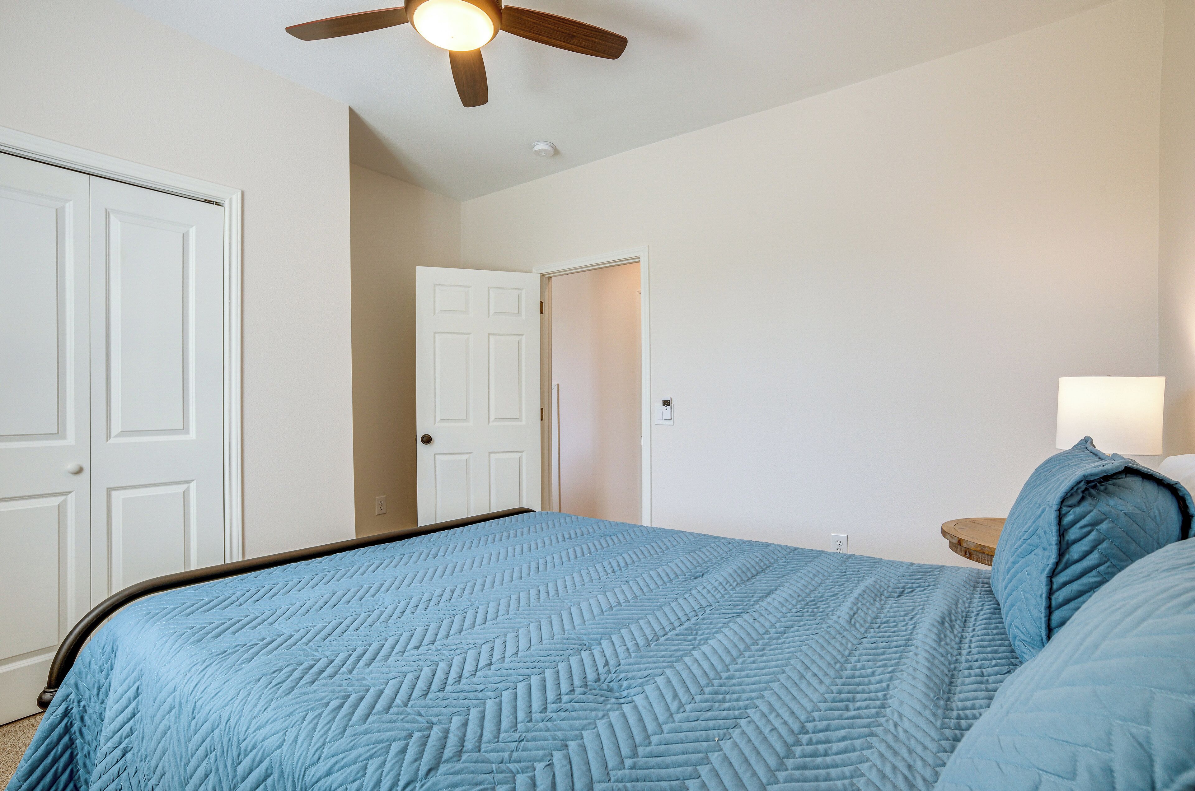 A bedroom with a bed, a ceiling fan, a nightstand, and a door leading to another room.