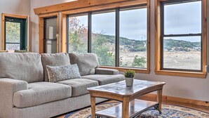 Una sala de estar con un sofá gris, una mesa de centro de madera y ventanas amplias que ofrecen vistas a un paisaje montañoso.