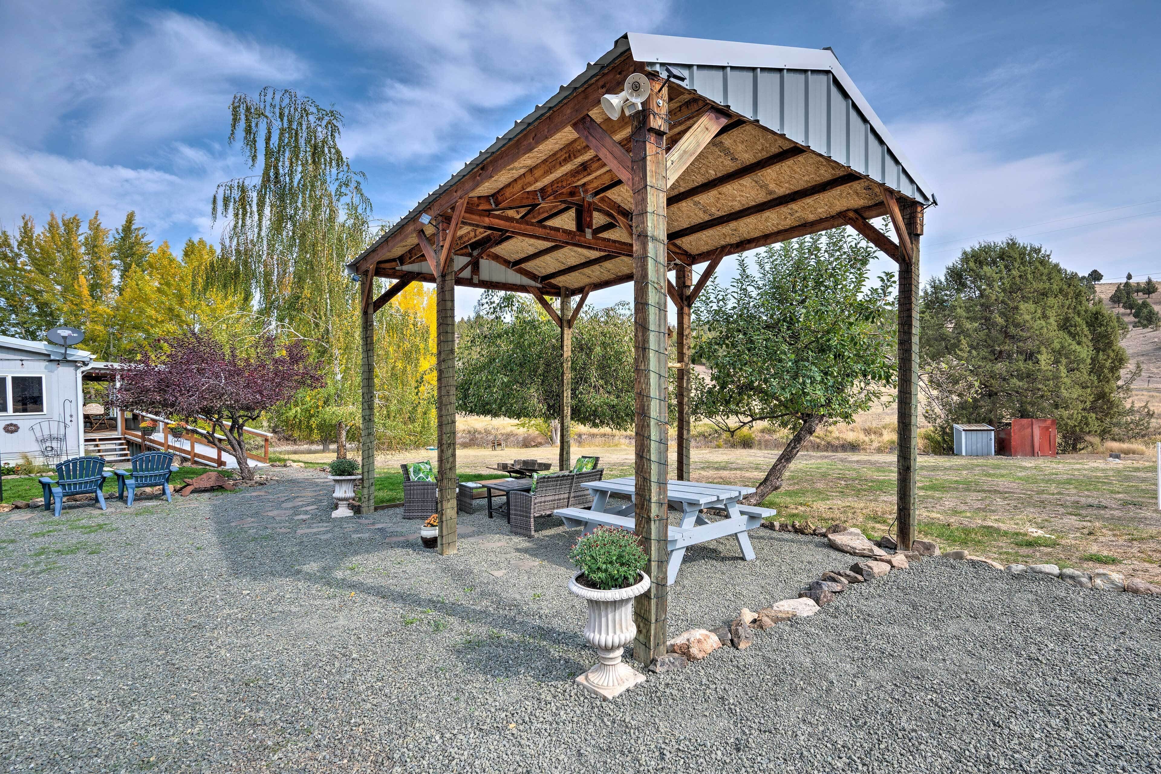 A covered outdoor seating area with picnic tables and chairs, surrounded by gravel and trees.