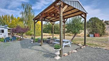 A covered outdoor seating area with picnic tables and chairs, surrounded by gravel and trees.