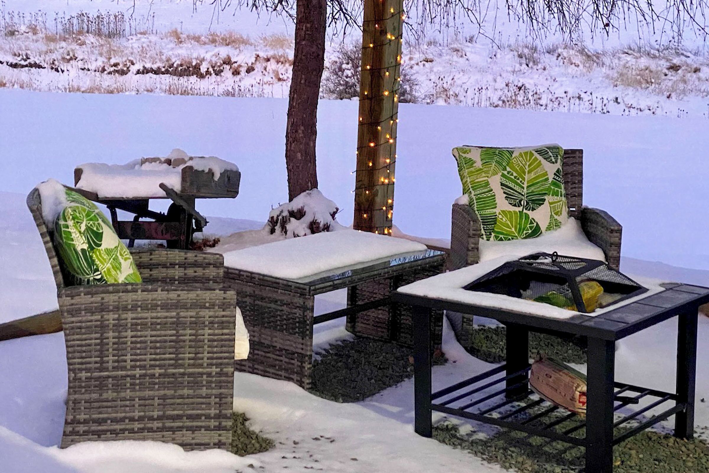 Outdoor wicker furniture with snow accumulation, a small table, and a tree with string lights.