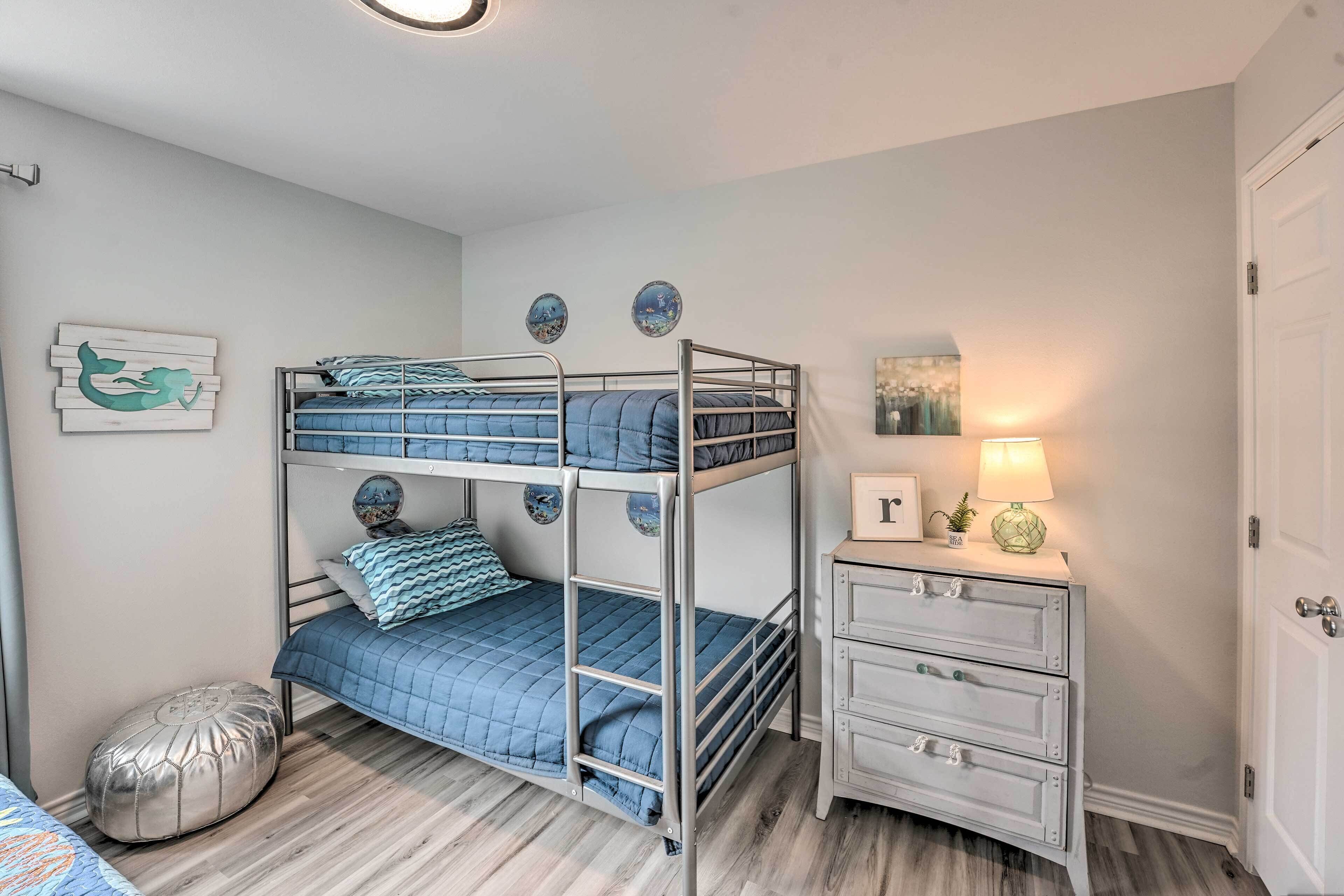 Un lit superposé avec une couette bleue, un petit pouf rond, une table de chevet avec une lampe et une commode blanche avec des tiroirs.